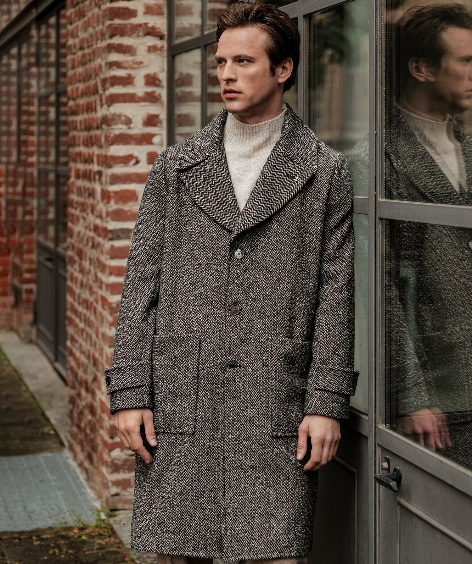 Man wearing a gray coat standing in front of a brick wall and glass door.