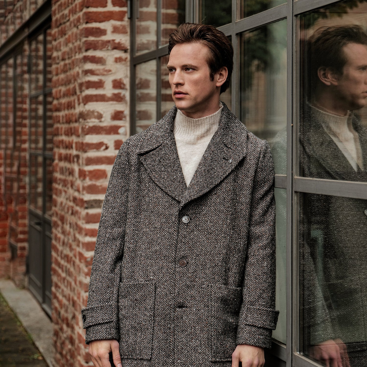 Man wearing a Distretto 12 grey coat standing in front of a brick wall and glass door.