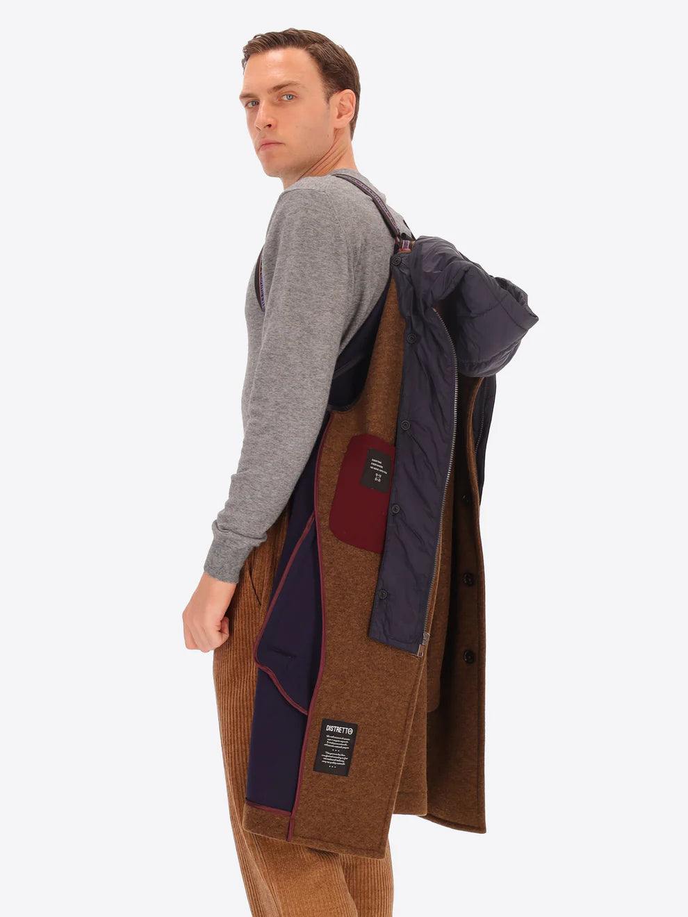 Male model wearing tobacco coloured overcoat by distretto12 with detachable hood hung over shoulder by straps
