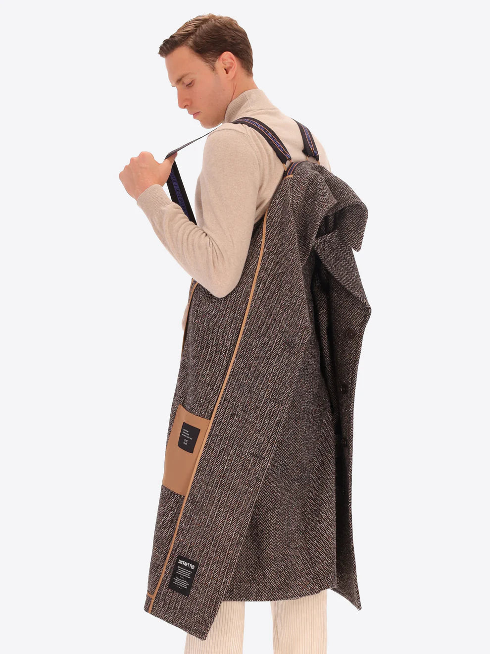 Male model wearing tobacco coloured overcoat by distretto12 with micro pattern hung over shoulder with back straps